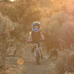 A child riding the Early Rider Seeker X16 16" Bike - Brushed Aluminium through shrubs and trees, on a dirt trail