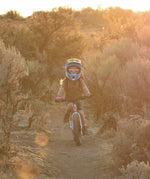 A child riding the Early Rider Seeker X16 16" Bike - Brushed Aluminium through shrubs and trees, on a dirt trail