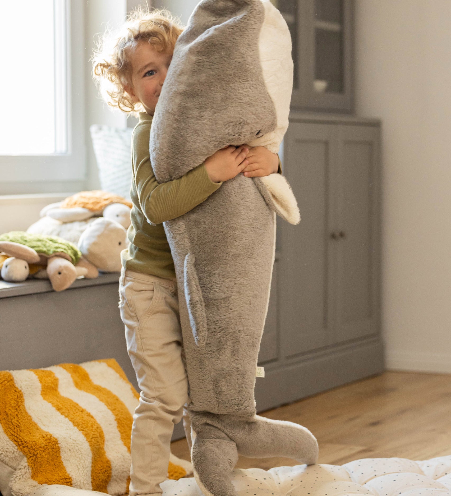 A child playing and holding the Senger Giant grey whale available at Babipur. A soft and cuddly giant whale toy in grey with playful facial features