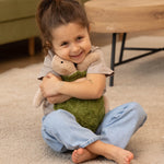 A child smiling happily and cuddling the Senger Cuddly Turtle - Green, whilst sitting down on a cream carpet/rug
