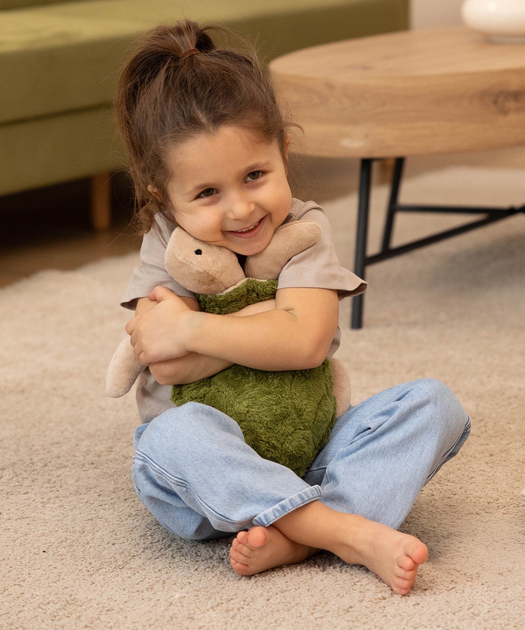A child smiling happily and cuddling the Senger Cuddly Turtle - Green, whilst sitting down on a cream carpet/rug