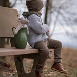 A child is sat on a light brown bench whilst looking off into the distance. The Senger Baby Mouse is sat inside a green ceramic jug, which shows the top half of the mouse. The jug is placed next to the child on the bench