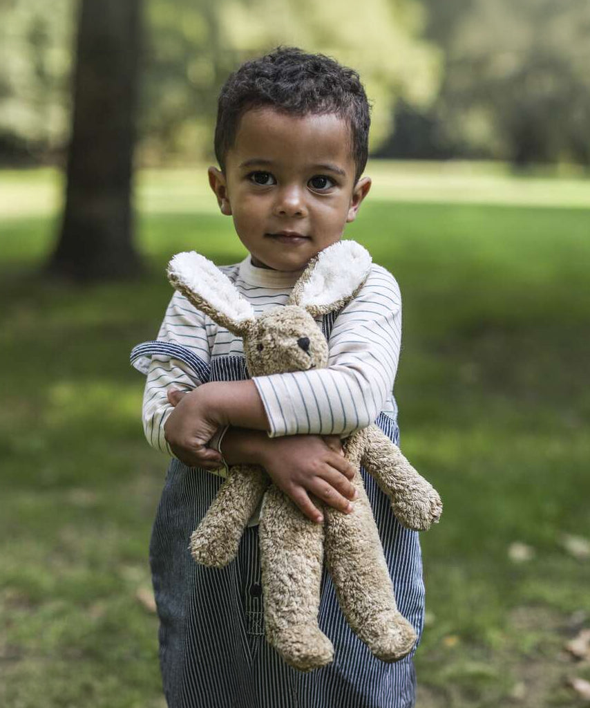 A child outside in a grassy field, holding the Senger Small Floppy Beige Rabbit in their arms