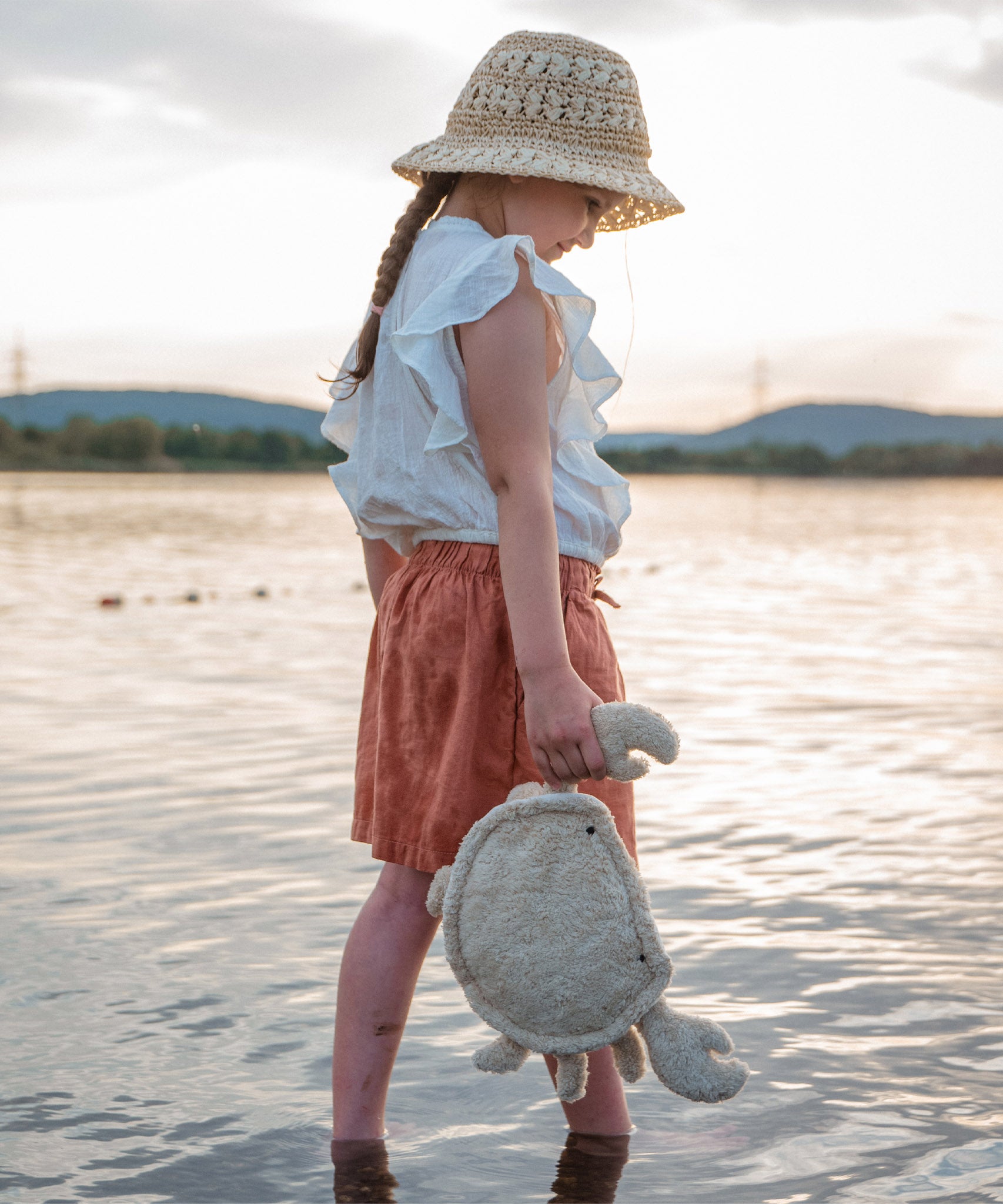 A small child standing in water and holding the cream coloured Senger Naturwelt small cuddly crab in a light beige colour by the claw