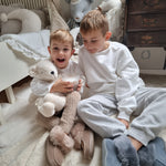 Two children sat on a white fluffy rug in their room. The child on the left is holding the Senger Large Cuddly Polarbear in their arms and a cup of hot chocolate