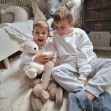 Two children sat on a white fluffy rug in their room. The child on the left is holding the Senger Large Cuddly Polarbear in their arms and a cup of hot chocolate