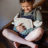 A small child sitting on wooden stairs and hugging the Senger Naturwelt small cuddly crab in a light beige colour with little black eyes, organic GOTS cotton plush soft toy with natural wool filling and spelt husk heat cushion.