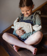 A small child sitting on wooden stairs and hugging the Senger Naturwelt small cuddly crab in a light beige colour with little black eyes, organic GOTS cotton plush soft toy with natural wool filling and spelt husk heat cushion.