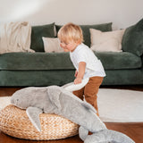 A small child playing and holding the fin of the grey Senger Naturwelt XL Cuddly Shark, organic GOTS cotton plush soft toy with removable spelt husk heat cushion.