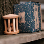 The Wooden Story Turtle Dove Rattle with its cardboard box next to is, is sat on a wooden plank next to a tree trunk