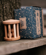 The Wooden Story Turtle Dove Rattle with its cardboard box next to is, is sat on a wooden plank next to a tree trunk