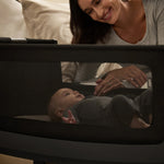 A woman watching over her baby from bed, with the SnuzPod5 close to her bedside. We see the baby through the black mesh of the SnuzPod.