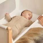 A small baby sleeping in the SnuzPod5 natural wooden bedside cot, in a cream babygrow.