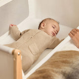A small baby sleeping in the SnuzPod5 natural wooden bedside cot, in a cream babygrow.