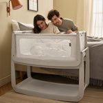 Two parents sat up in bed, watching their baby as it lays beside them in the 'Pebbles' coloured SnuzPod5 bedside crib.
