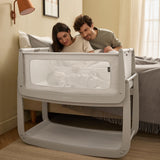 Two parents sat up in bed, watching their baby as it lays beside them in the 'Pebbles' coloured SnuzPod5 bedside crib.