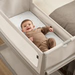 A small baby laying in the SnuzPod5 pebbles grey wooden bedside cot, in a light brown babygrow.