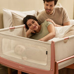 Two parents watch their baby from bed, the baby is in a Terracotta SnuzPod5 bedside cot next to them.