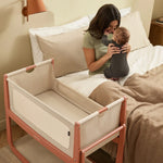 A woman sat in bed holding her baby, with the SnuzPod5 in terracotta right beside the bed. 