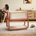 The Terracotta wooden SnuzPod5 bedside crib with wheels, that has been wheeled into the living room whilst a mother holds her baby. 