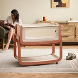 The Terracotta wooden SnuzPod5 bedside crib with wheels, that has been wheeled into the living room whilst a mother holds her baby. 