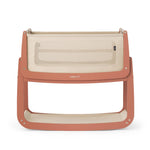 The SnuzPod5 bedside baby crib with a terracotta pink wooden frame, cream mesh sides, and a cream mattress. 