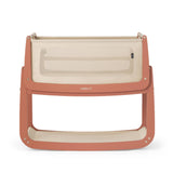The SnuzPod5 bedside baby crib with a terracotta pink wooden frame, cream mesh sides, and a cream mattress. 