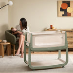 The sage wooden SnuzPod5 bedside crib with wheels, that has been wheeled into the living room whilst a mother holds her baby. 