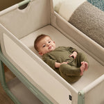 A small baby laying in the SnuzPod5 sage green wooden bedside cot, in a green babygrow.