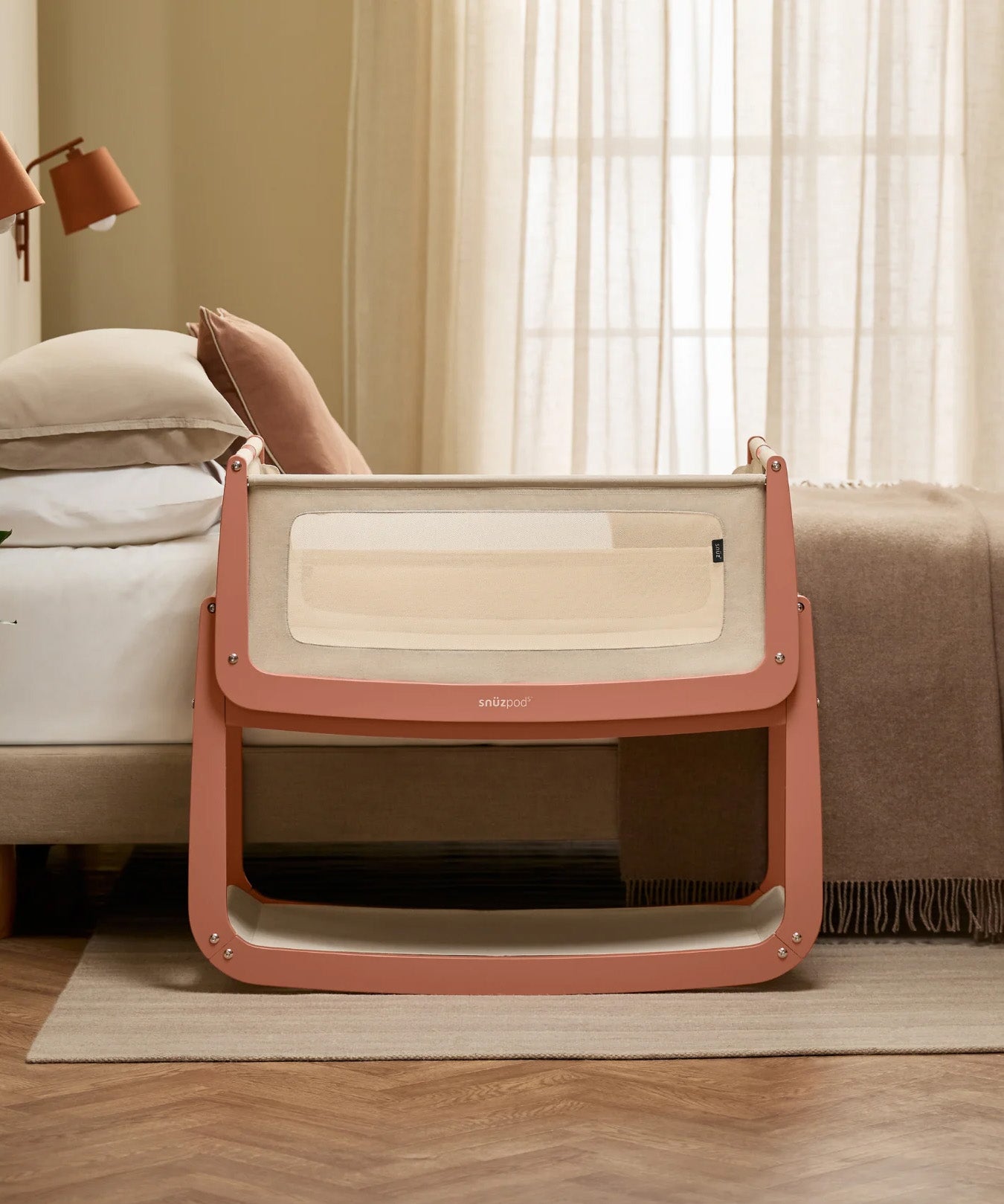 The SnuzPod 5 bedside baby crib with a lovely terracotta pink wooden frame, next to an adult's bed in a beautifully decorated bedroom.