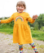 A child happily wearing the Frugi Niamh Dress - Gold/Rainbow. The dress is paired with the Frugi Retro Happy Norah Tights