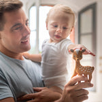 A child being held by an adult, whilst reaching out towards the Fanfan The Fawn Baby Teether, being held in the parents hand