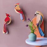 The Studio Roof Jay and Cardinal birds are on a dark pink, textured background. On the right hand side of the image sits the Jay, with a blue shelf underneath the model and a small blue vase with purple and green flowers.