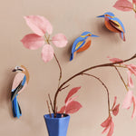 The Studio Roof Kingfisher and Jay birds on a peach wall, with a pink plant in a blue vase, at the foreground of the image