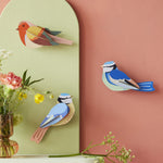 The Studio Roof Blue Tits and Robin bird, on a pink and green background, with flowers laid on a white shelf underneath the birds