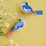 The Studio Roof Blue Tits Cardboard bird Modelon a yeallow background, with daisy flowers on the left hand side of the photo