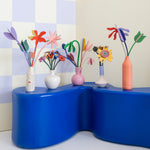 A collection of Studio Roof cardboard flowers in colourful vases, stood on a blue table