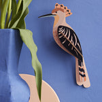 The Studio Roof Hoopoe Cardboard Model, set on a blue coloured background. On the left hand side of the image is a blue vase with a green plant inside.