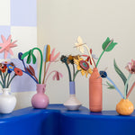 A collection of Studio Roof cardboard flowers in colourful vases, stood on a wooden table
