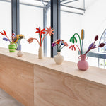 A collection of Studio Roof cardboard flowers in colourful vases, stood on a wooden table