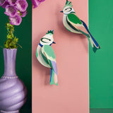 Studio Roof forest crests wall decorations available at Babipur. Hanging on a pink board against a green wall with a purple vase in front.