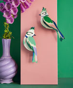 Studio Roof forest crests wall decorations available at Babipur. Hanging on a pink board against a green wall with a purple vase in front.