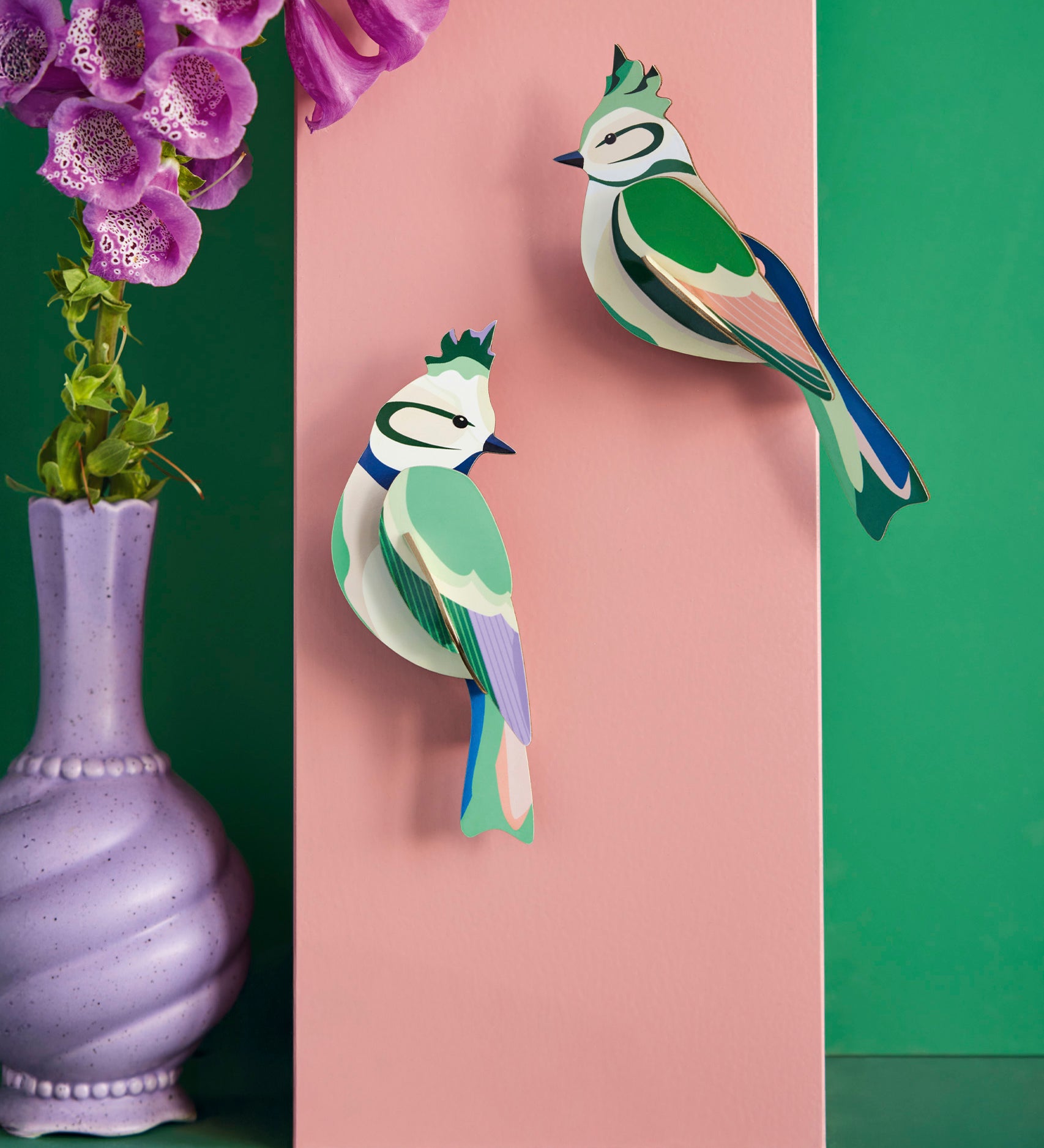 Studio Roof forest crests wall decorations available at Babipur. Hanging on a pink board against a green wall with a purple vase in front.