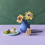 The Studio Roof Cardboard Bouquet - Van Gogh Sunflowers, stood in a blue vase. In front of the vase is one of the sunflwers laying horizontally and next to the vase on the left hand side is a small, light green plate with a lemon a a stripy cup. The image is on a blue and lilac background
