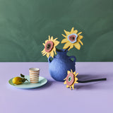 The Studio Roof Cardboard Bouquet - Van Gogh Sunflowers, stood in a blue vase. In front of the vase is one of the sunflwers laying horizontally and next to the vase on the left hand side is a small, light green plate with a lemon a a stripy cup. The image is on a blue and lilac background