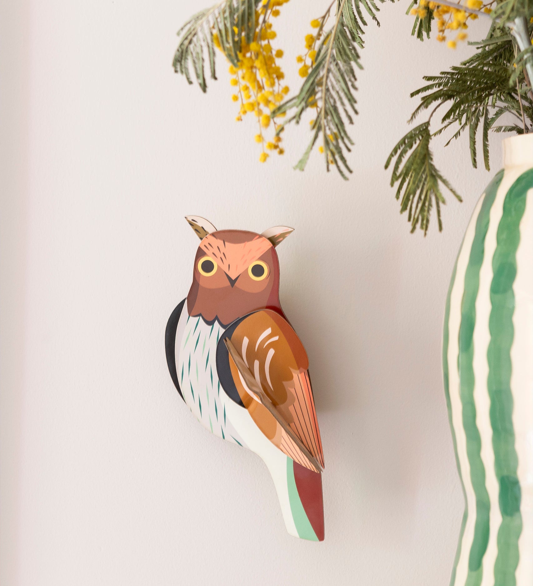 Studio Roof tawny owl wall decoration available at Babipur. Hanging on a lightly coloured wall with a striped vase in front.