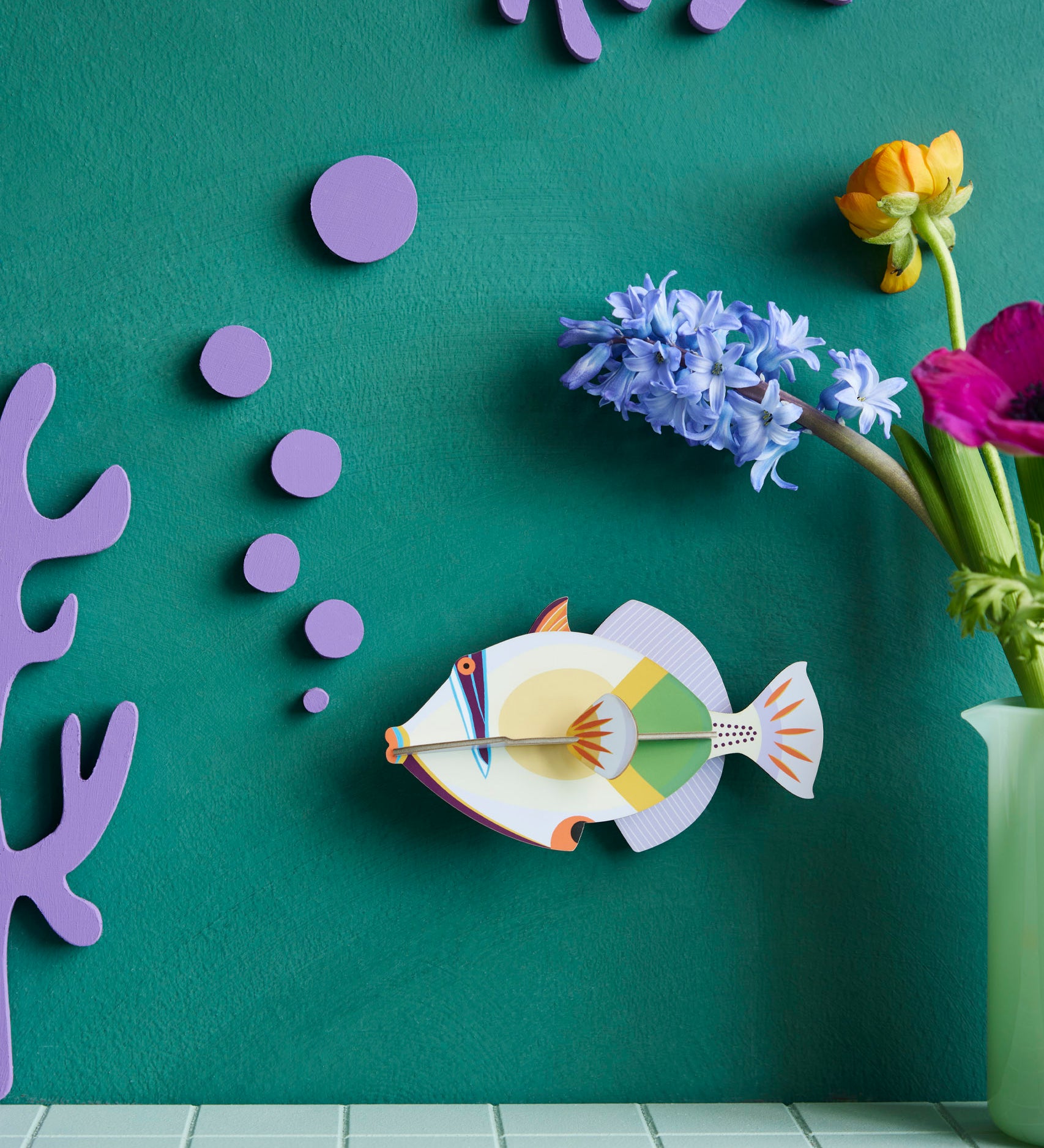 Studio Roof lagoon triggerfish wall decoration available at Babipur hanging on a green wall next to a green vase