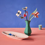 The Studio Roof Cardboard Bouquet - William Morris in a painted green vase, with a cream painted bowl with little papermache balls inside, and a green and white stripe cardboard cup in the background. The image is on a blue/purple and peach background