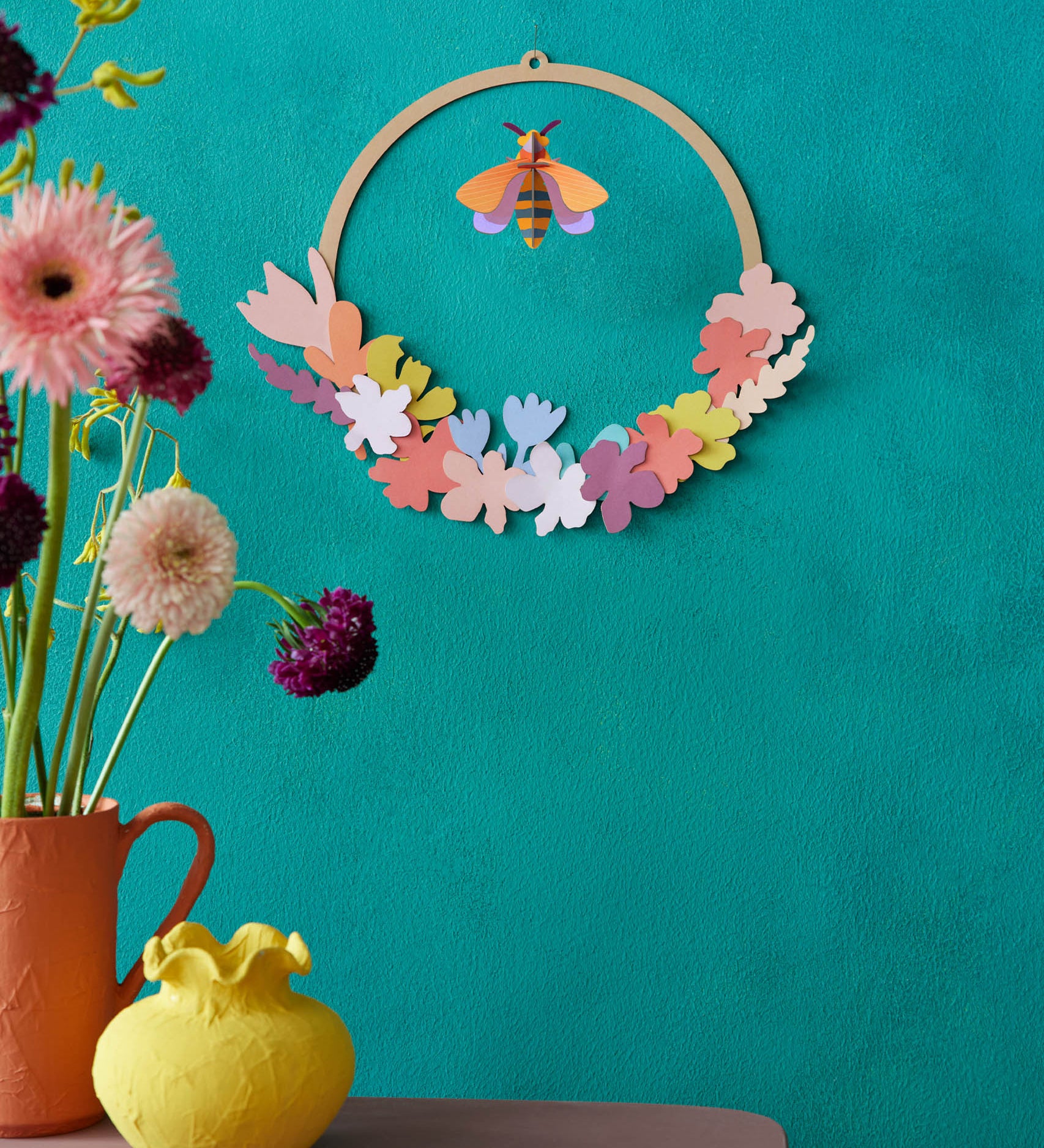 A decorative display with Studio Roof wreath meadow with bee available at Babipur. A DIY kit made from cardboard thats hanging on a wall