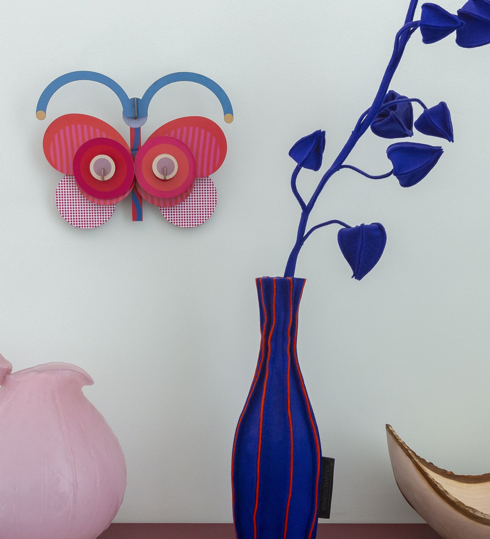 Stripy butterfly wall decoration available at Babipur hanging on a wall above a pink and blue vase. A Studio roof collaboration with artist Zoe Miller. 